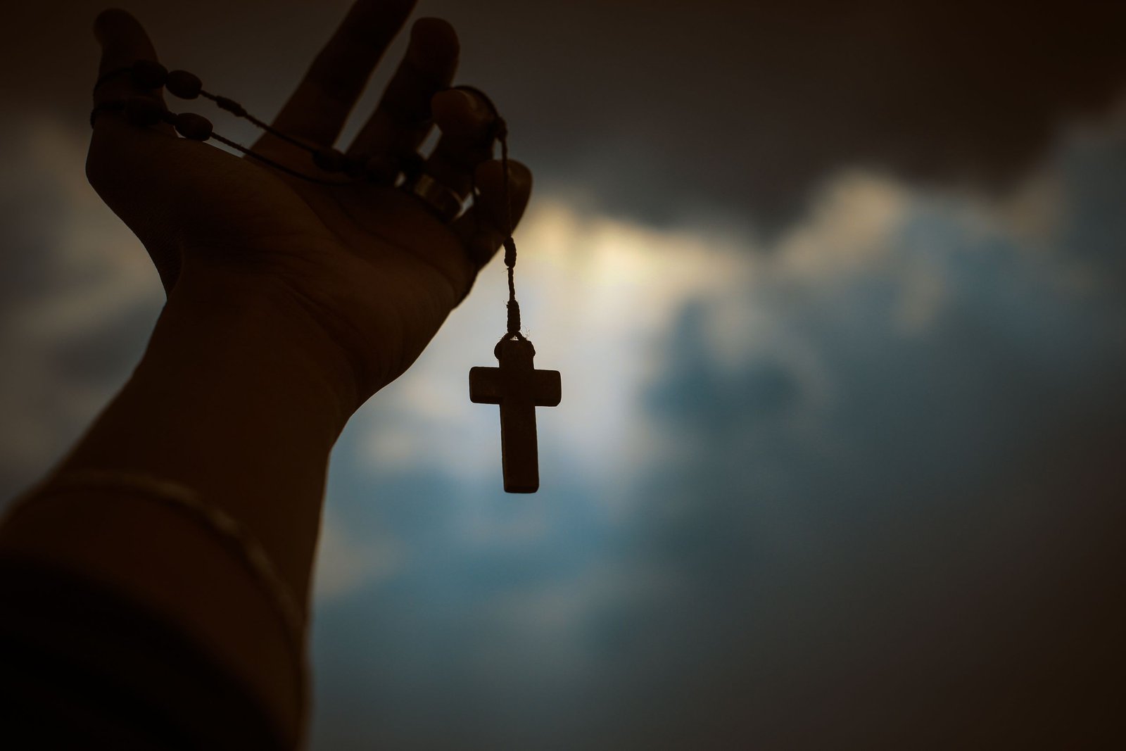 Silhouette of a hand holding a rosary with a cross against a dramatic sky, symbolizing faith and spirituality.